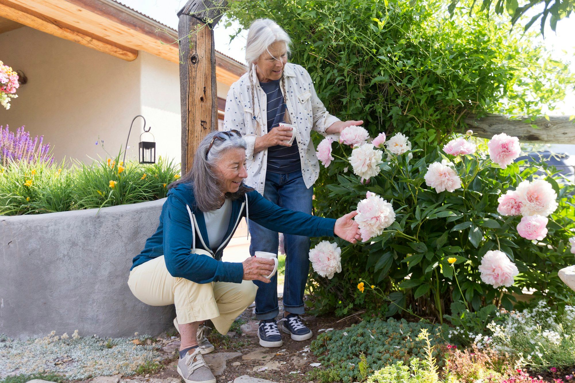 Two older women enjoying pink peonies in a sunny garden